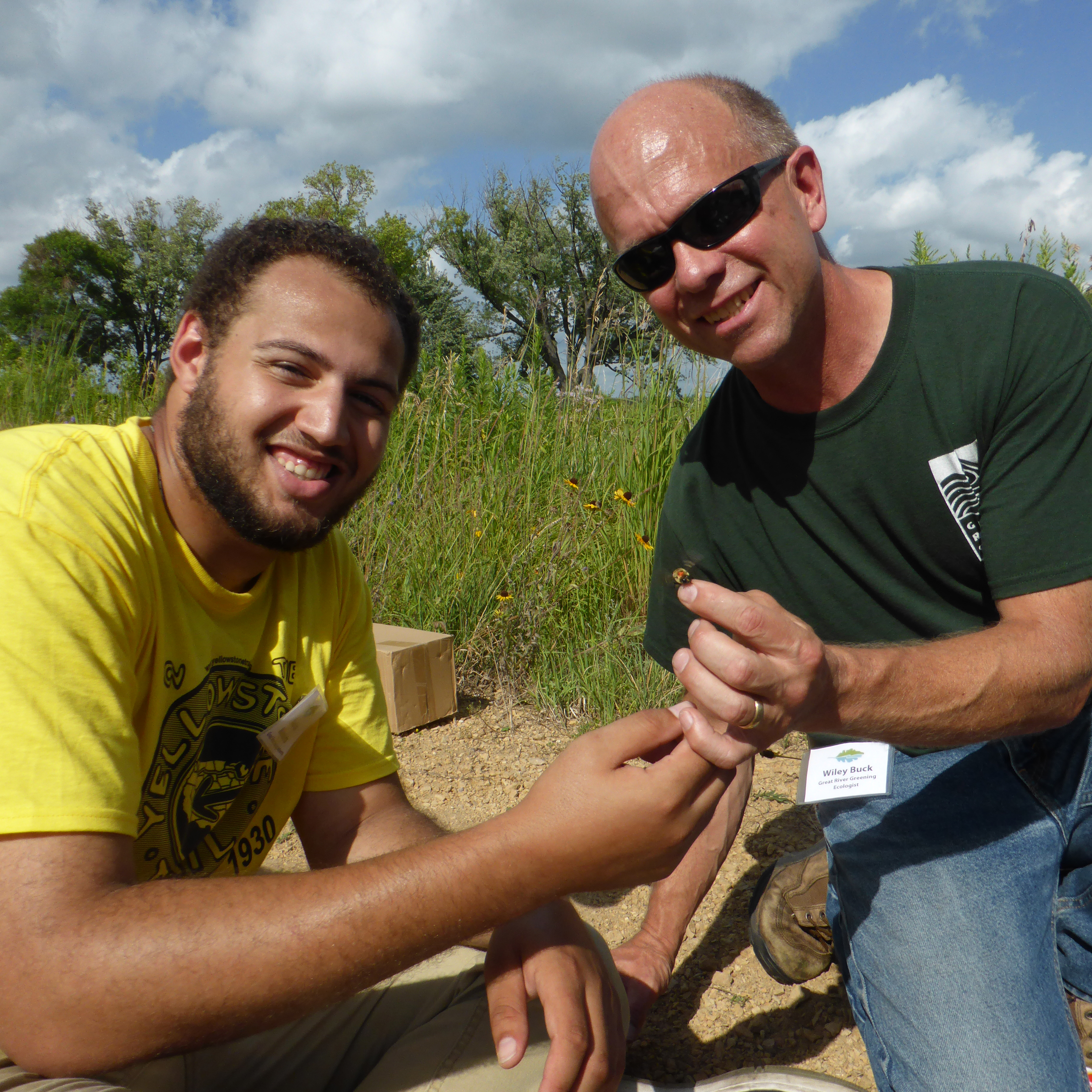 Two people, one a young man with dark skin, and one an older man with pale skin, have their hands close together as they show off a bumble bee they caught as part of a community science effort.