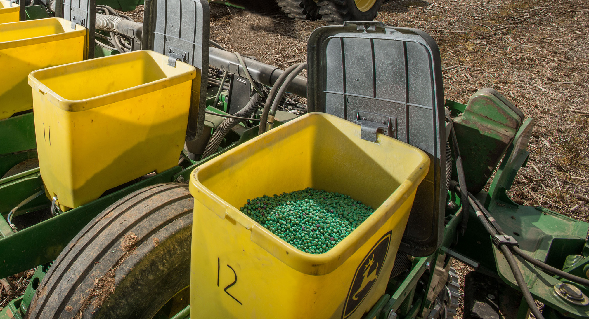 "A close-up of a seed drill, a large piece of farming equipment towed behind a tractor to sow seed. The metal frame of the seed drill is green and grubby. The tire has dried mud caked on it. Several rectangular yellow plastic bins on the drill are full of seed corn. The seed corn is green and pink."