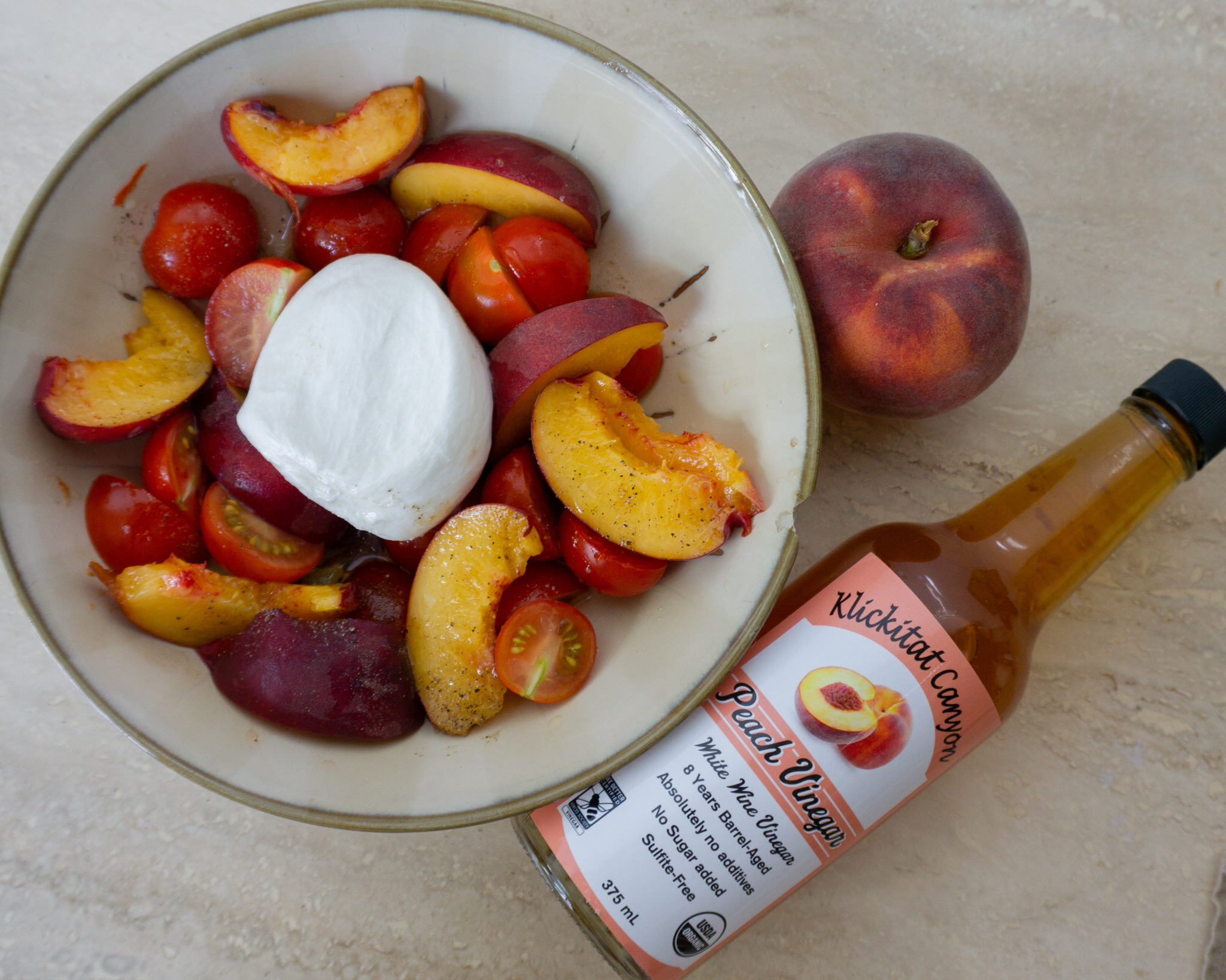 A bottle of vinegar lies on a table beside a bowl full of slices of peach and cherry tomatoes