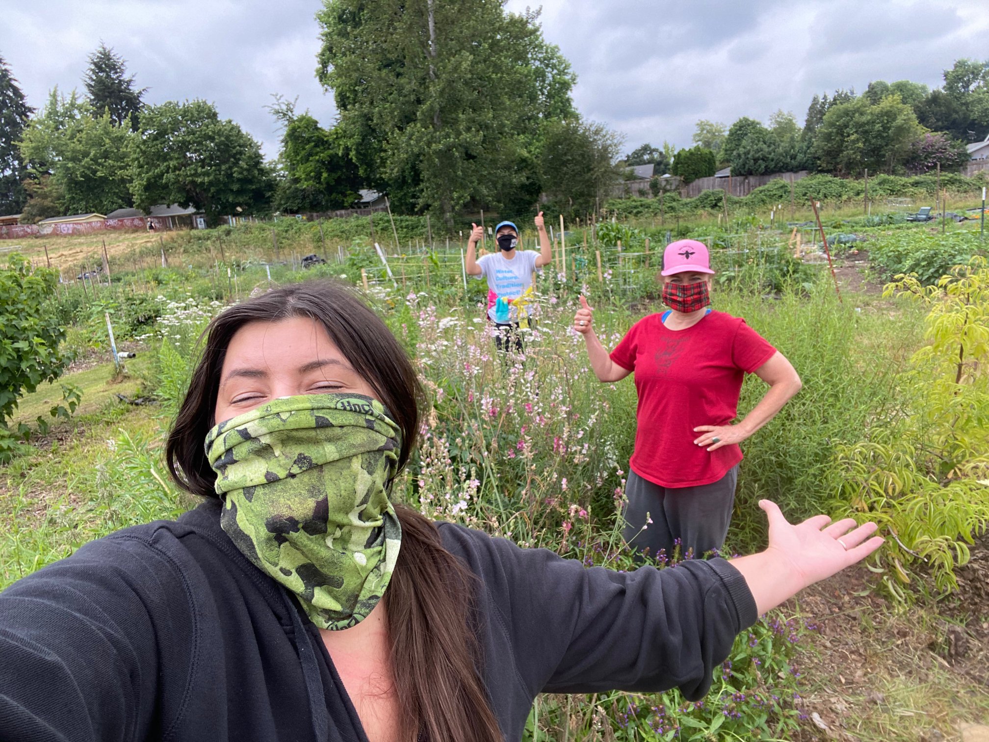A self-portrait of a woman with long dark hair and wearing a green pattern mask. She is gesturing toward two other women working with her in a community garden.