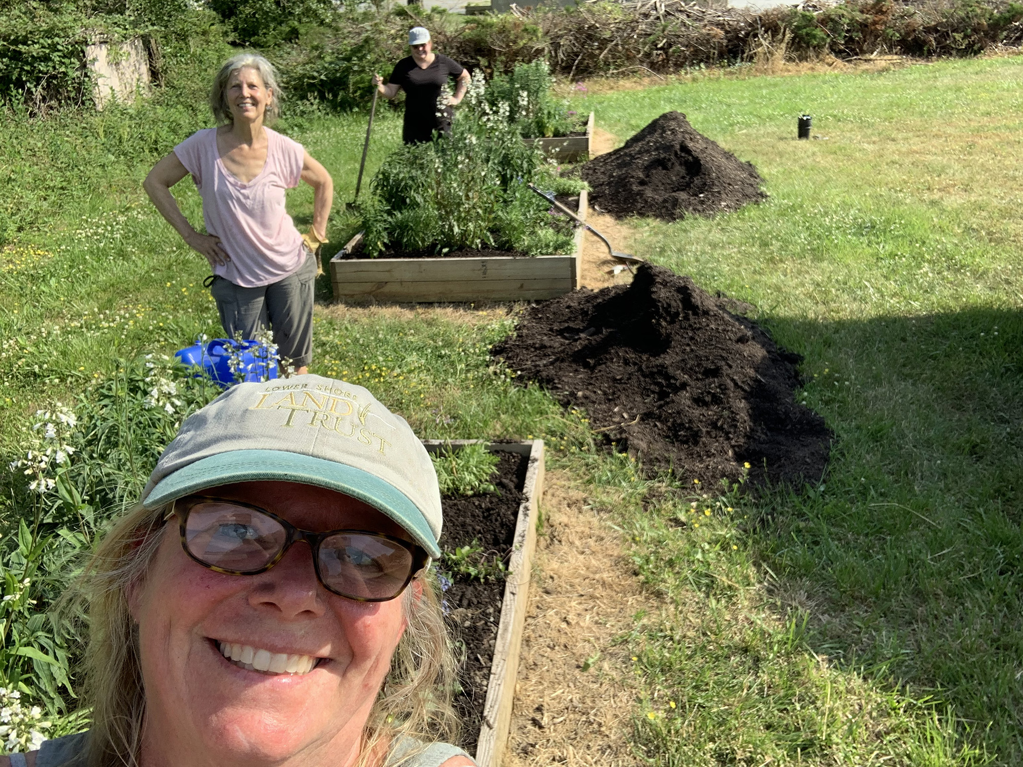 A self portrait of a woman wearing a green cap and with brown-rimmed glasses. Be hind her are two women smiling at the camera beside the raise beds they have been working to plant.