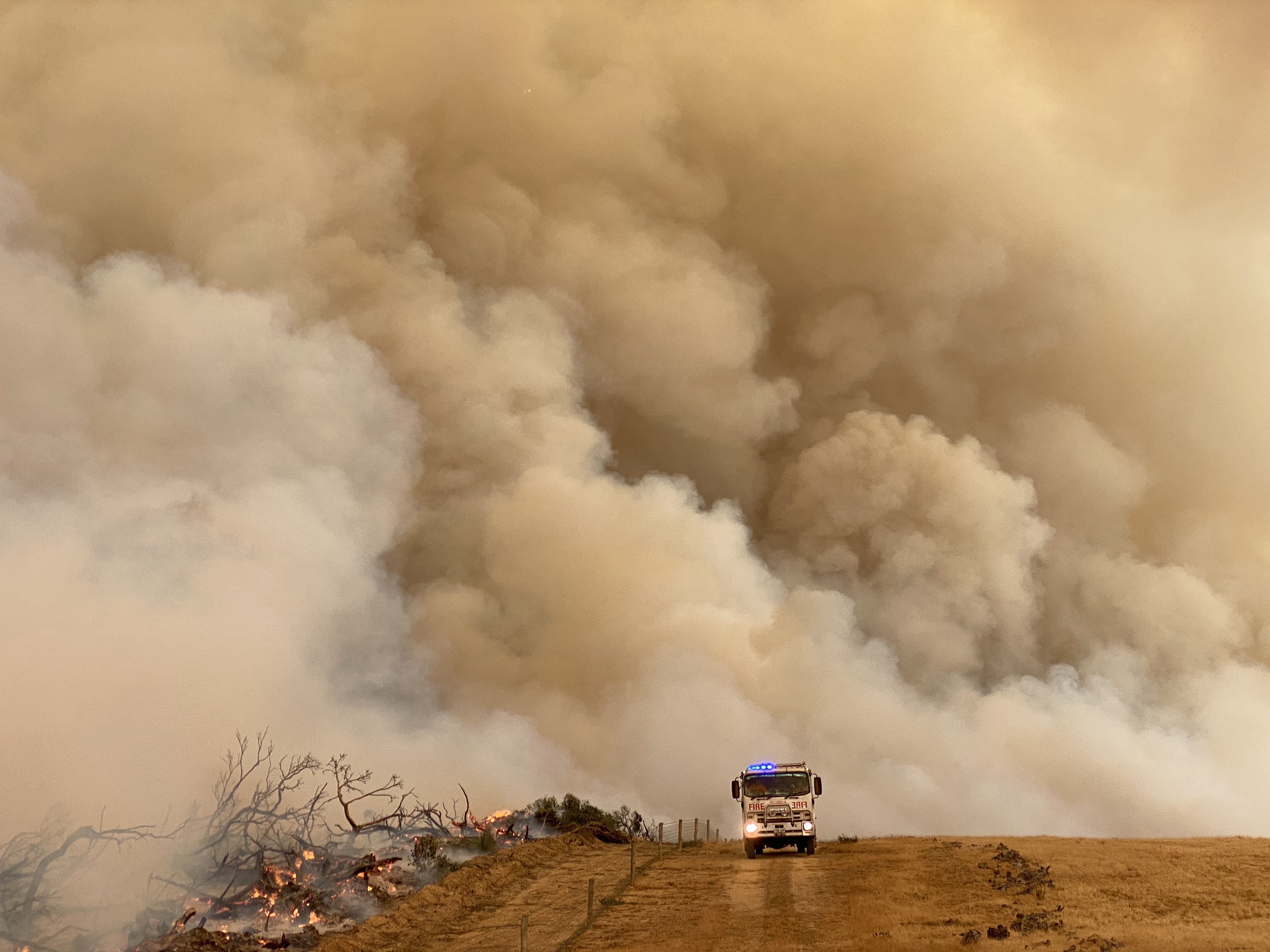 Fire truck drives across a burning landscape, with the sky full of brown smoke