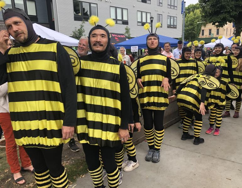 A long line of people of diverse ages, genders, and races are all sporting bright yellow and black-striped bee outfits in an outdoor festival setting.