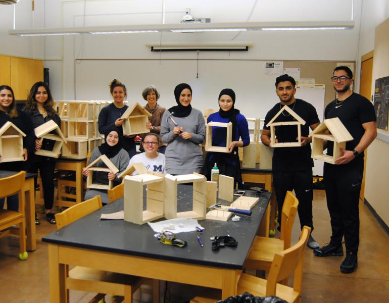 A group of young people standing a classroom holding wooden insect houses that they have been building. Some people have blonde hair, some brown, and some black. A few of the group are wearing head scarfs. The insect houses are square frames with a pointed roof.