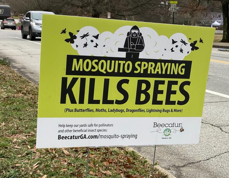 A roadside sign that is a bright green color with the message “MOSQUITO SPRAYING KILLS BEES” in black text. A silver-colored car and a bus drive past in the background.
