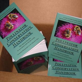 A brown cardboard box has been opened to show the contents, bundles of newly printed books. The books are a gray-green color, with a photograph that shows two yellow and orange bumble bees on a pink flower.