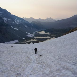 A Xerces conservation biologist hikes down from a small glacier that is now mostly just a snowfield. A chain of meltwater lakes can be seen in the distance, with smoke from nearby wildfires on the horizon.