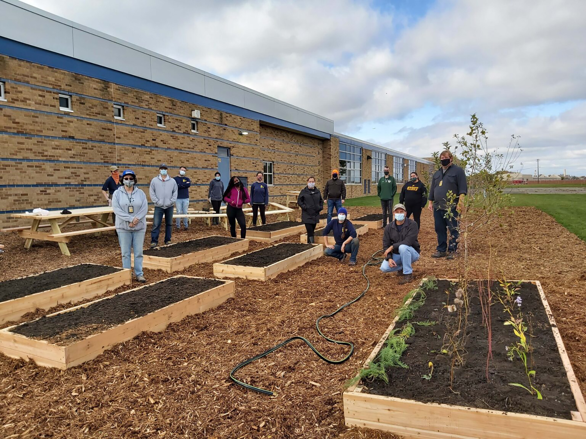 A group of people stand in the school garden that they have just finish building. They are wrapped against the cold day in green, black, dark blue, dark gray, pale gray, and red sweatshirts or jackets. The garden has several rectangular raised beds made with pale brown wood and filled with dark brown soil. Rows of green and red plants fill one of them. The beds are surrounds by wood chips and behind is the brick wall of the school building.