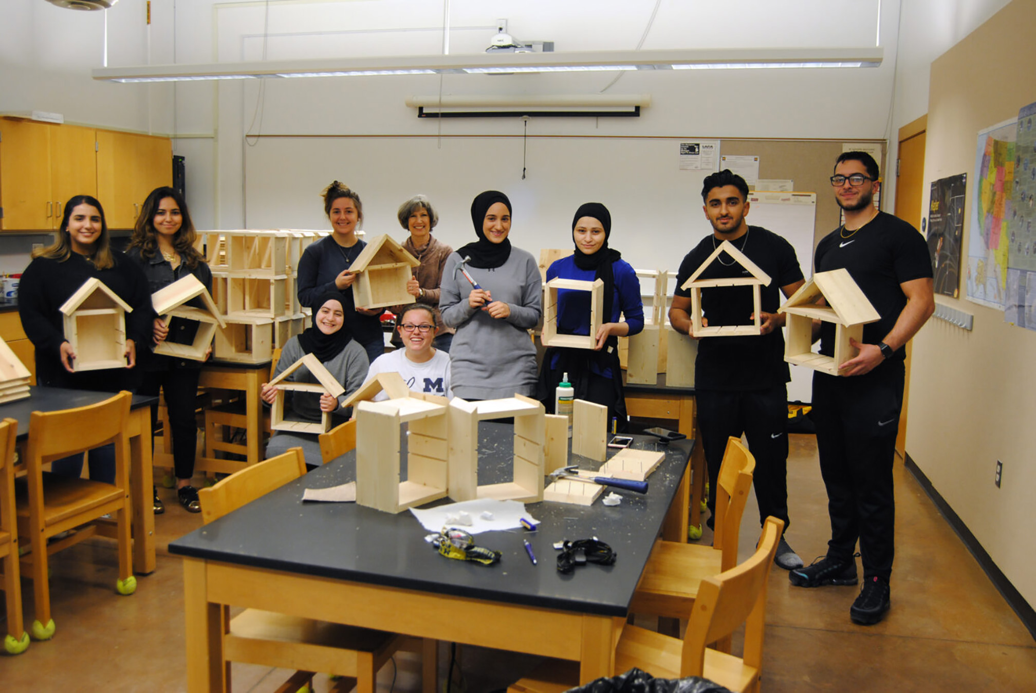 A group of young people standing a classroom holding wooden insect houses that they have been building. Some people have blonde hair, some brown, and some black. A few of the group are wearing head scarfs. The insect houses are square frames with a pointed roof.