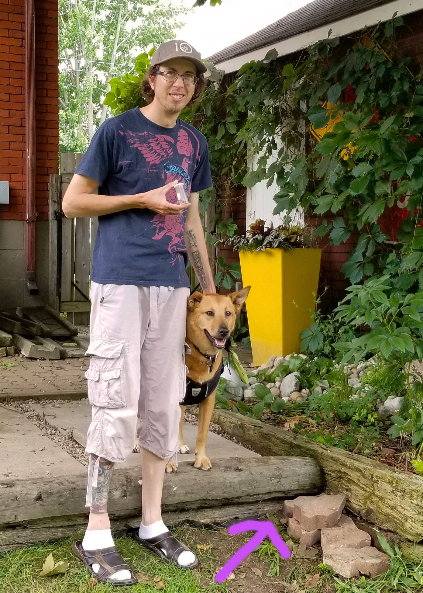A thin man with tattoos on his arms and legs stands, smiling, with a leashed dog. The man is holding a small, plastic tube in one hand. There is a drawn-in pink arrow pointing to a cluster of pavers on the grass in front of him and his dog.