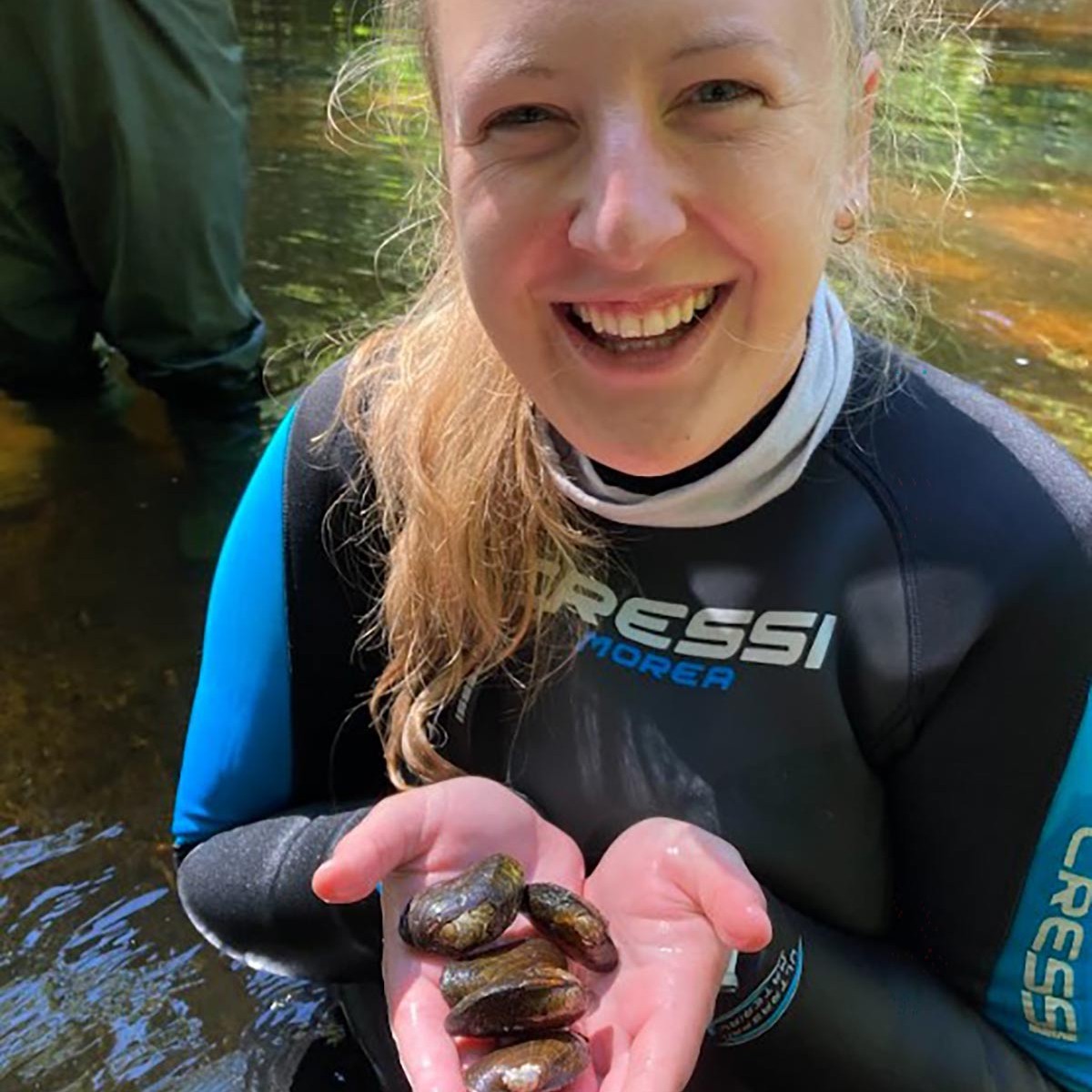 Smiling person in a wetsuit standing in water and holding several freshwater mussels in their hands