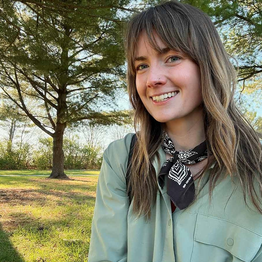 A smiling person sits outdoors in a sunny grassy area with trees in the background, wearing a light green shirt and black patterned neck scarf