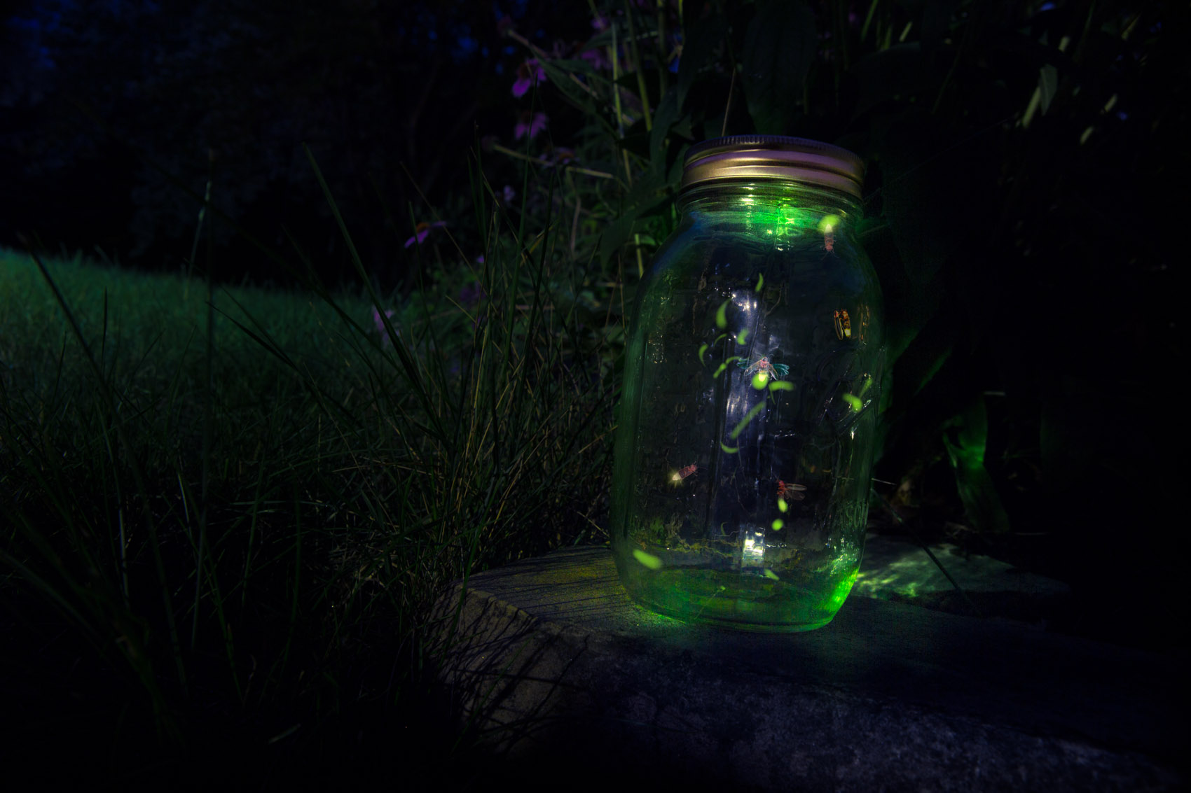 A glowing green mason jar contains a few fireflies in flight.