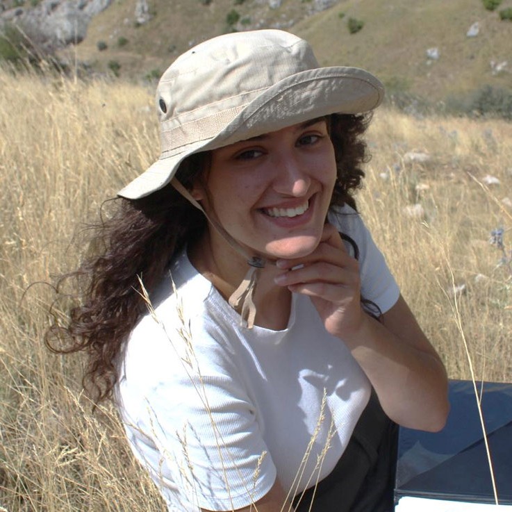 A smiling person wearing a wide-brimmed sun hat sits in a dry grassy hillside landscape with rocky hills in the background