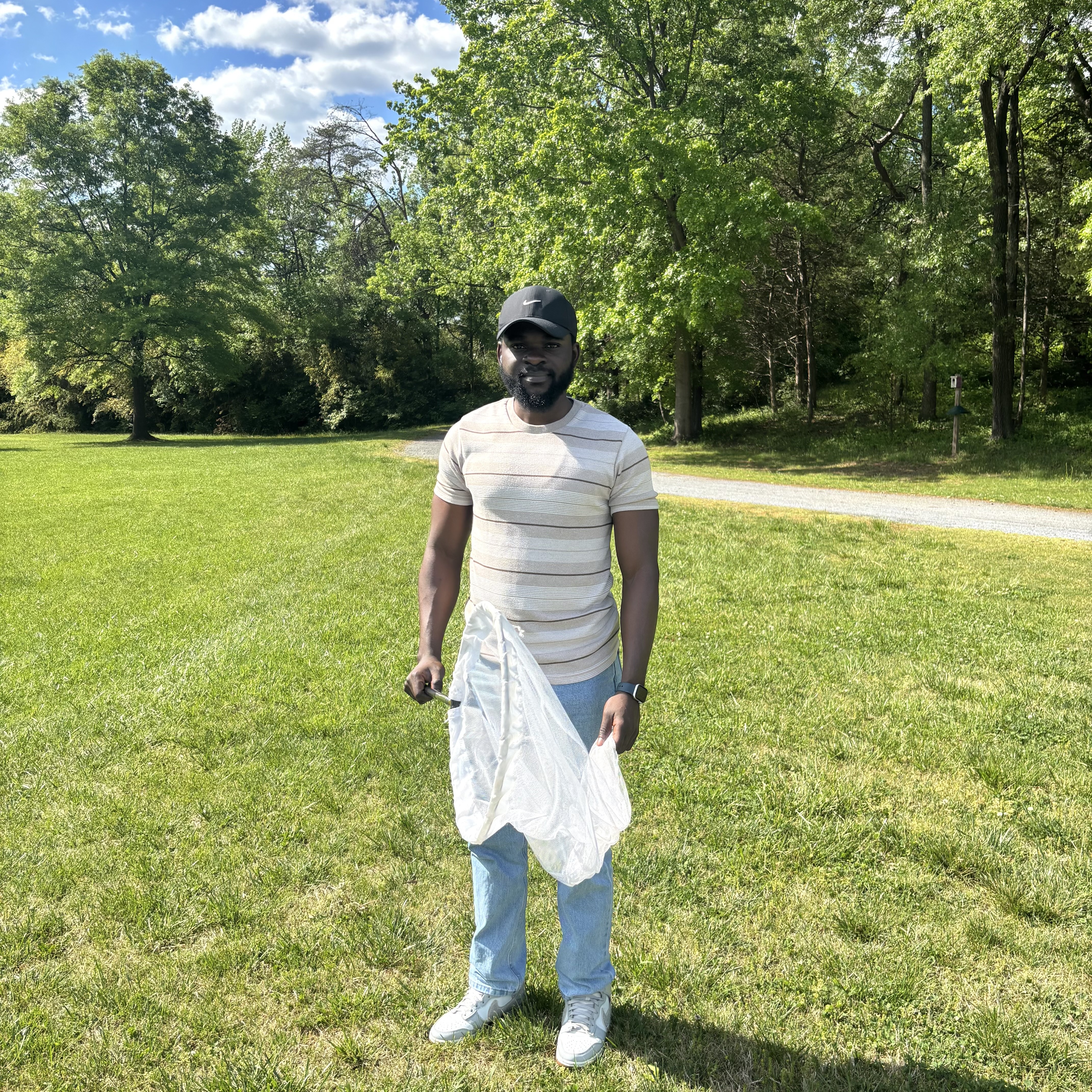 Man standing in a grassy park holding a white insect net, with trees and a path in the background
