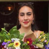 Person with dark hair and earrings holding a colorful bouquet of flowers, standing in a doorway and looking at the camera