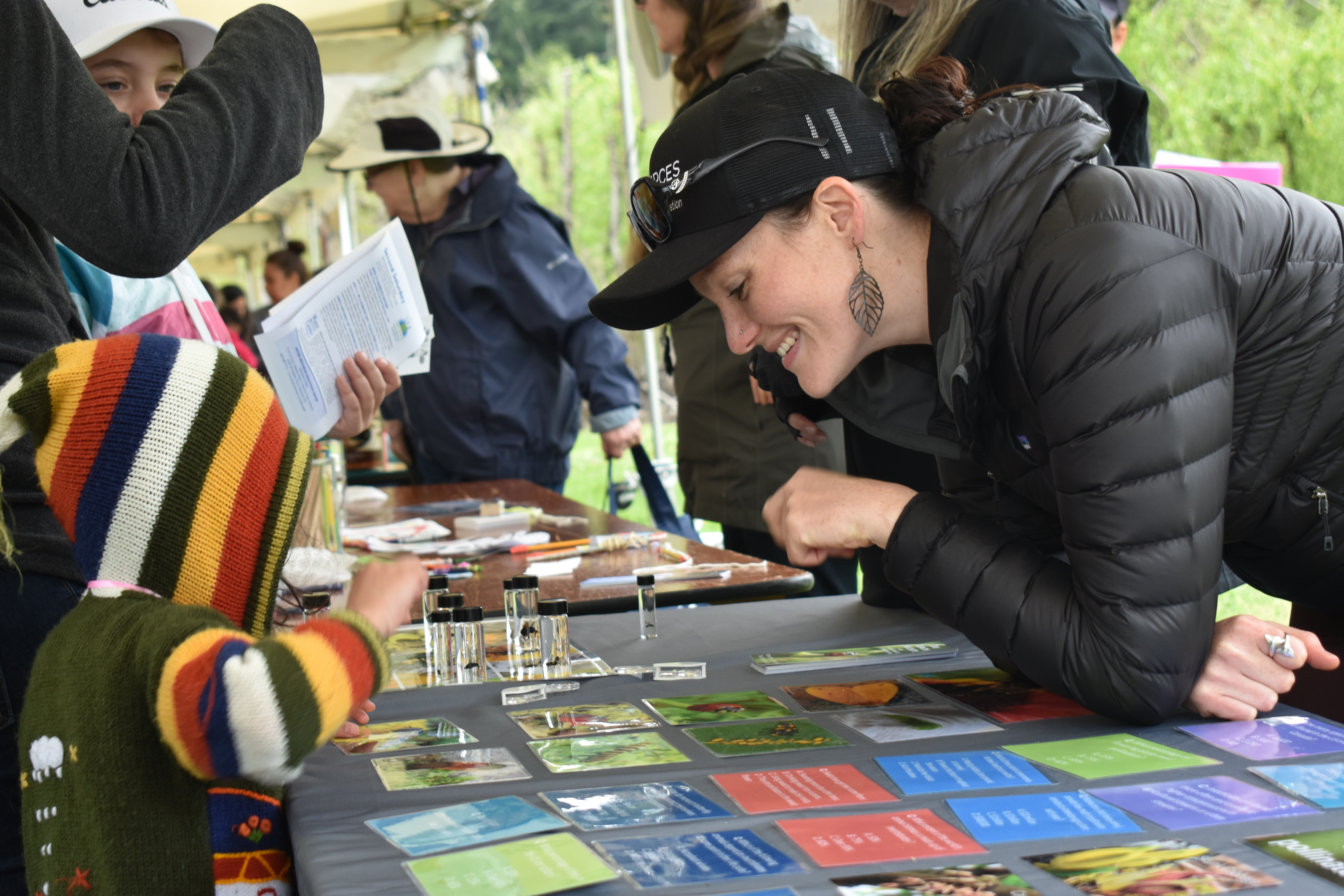 A smiling woman wearing a black coat and a black Xerces Society hat leans over a table. On the other side of the table, a kid with a colorful hoodie reaches for an assortment of small glass vials containing bee specimens. The woman and the kid are looking at the same vials and appear to be talking.