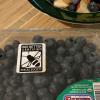 A clear plastic container of blueberries on a kitchen counter beside a breakfast bowl containing cereal and fruit. The blueberry container has the black-and-white Bee Better Certified label