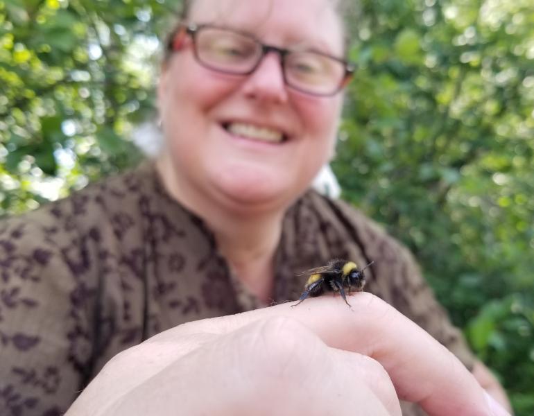 A woman smiles while looking at a fuzzy, black, and yellow bumble bee on her finger.