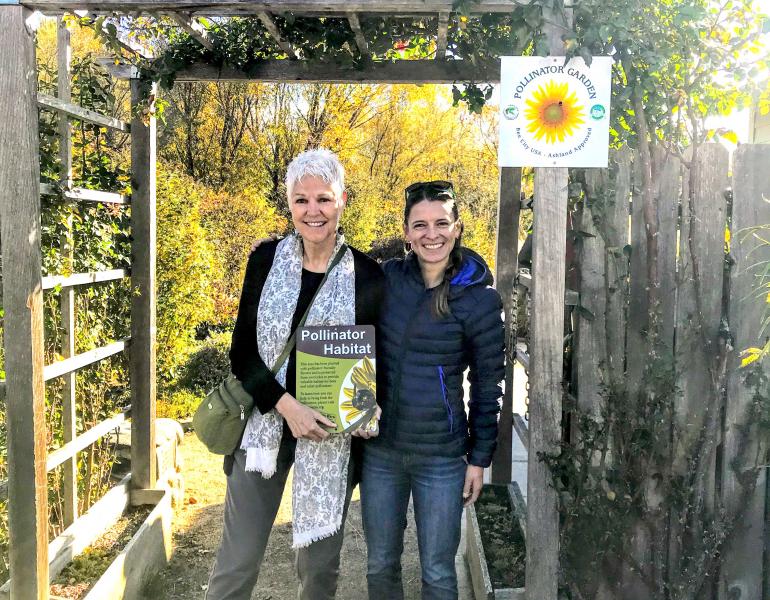 A tall woman with short, white hair who is holding a Xerces pollinator habitat sign stands next to a woman with dark hair and a dark down coat, in the entrance to a garden.