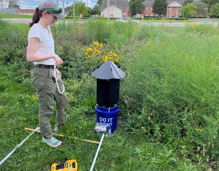 A white woman standing in a wildflower field, next to a set of equipment for collecting moths and surveying plant species. In the background, a road and houses show that this is in an urban setting.