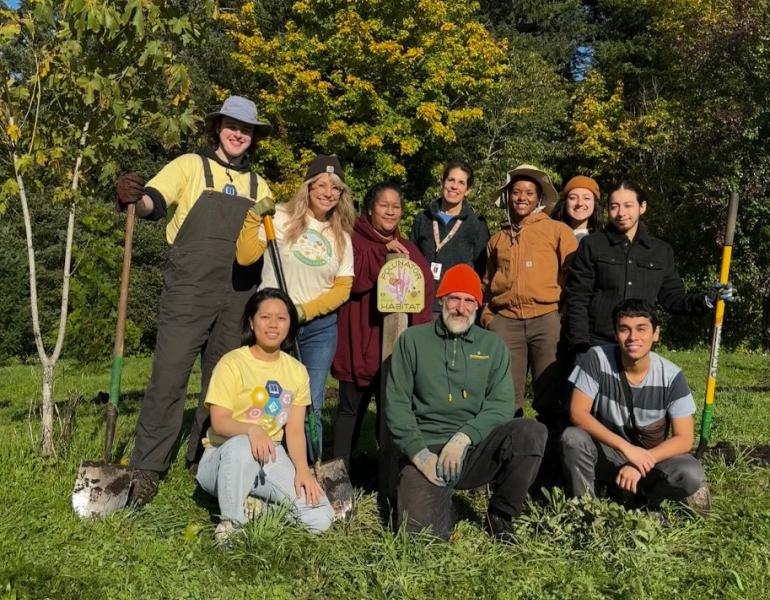 10 people in work clothes with shovels gather around a pollinator habitat sign in a garden.