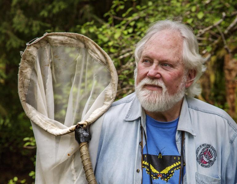 Robert Michael Pyle, who founded the Xerces Society in 1971. He has grey hair and beard, and is wearing a blue denim shirt over a blue t-shirt with a black-and-yellow swallowtail butterfly design -- and is holding his butterfly net.