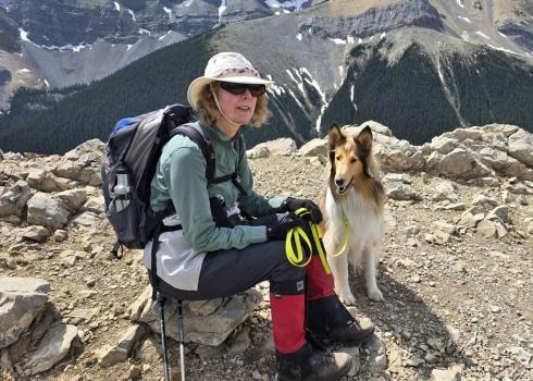 woman in hiking gear sits near her collie dog on top of a mountain summit