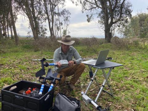 Researcher wearing a wide-brimmed hat kneels in a grassy field, assembling monitoring equipment beside a laptop on a small table