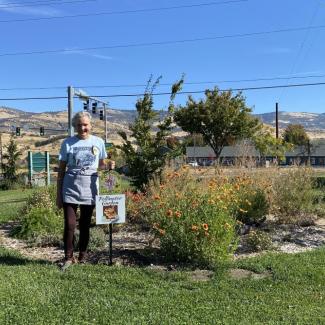 A woman with short, gray hair stands smiling in front of blooming native flowers and a pollinator habitat sign. Blue sky, power lines and hills are in the background.