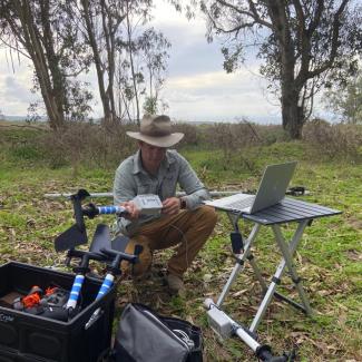 Researcher wearing a wide-brimmed hat kneels in a grassy field, assembling monitoring equipment beside a laptop on a small table