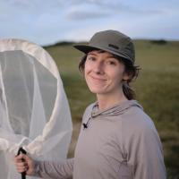 A person wearing a light gray long-sleeve shirt and an olive-green cap stands outdoors in a grassy landscape, smiling gently at the camera while holding a large white insect net.