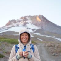 Smiling person in a hooded fleece holding a mug on a rocky mountain trail, with a snow-covered peak in the background