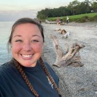 Smiling person taking a selfie on a rocky beach near calm water, with driftwood, grassy shoreline, and a few people in the background