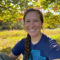 A smiling woman with a dark braid and orange sunglasses on her head sits outdoors in front of a sunlit autumn meadow and colorful foliage, wearing a navy Lake Superior t-shirt